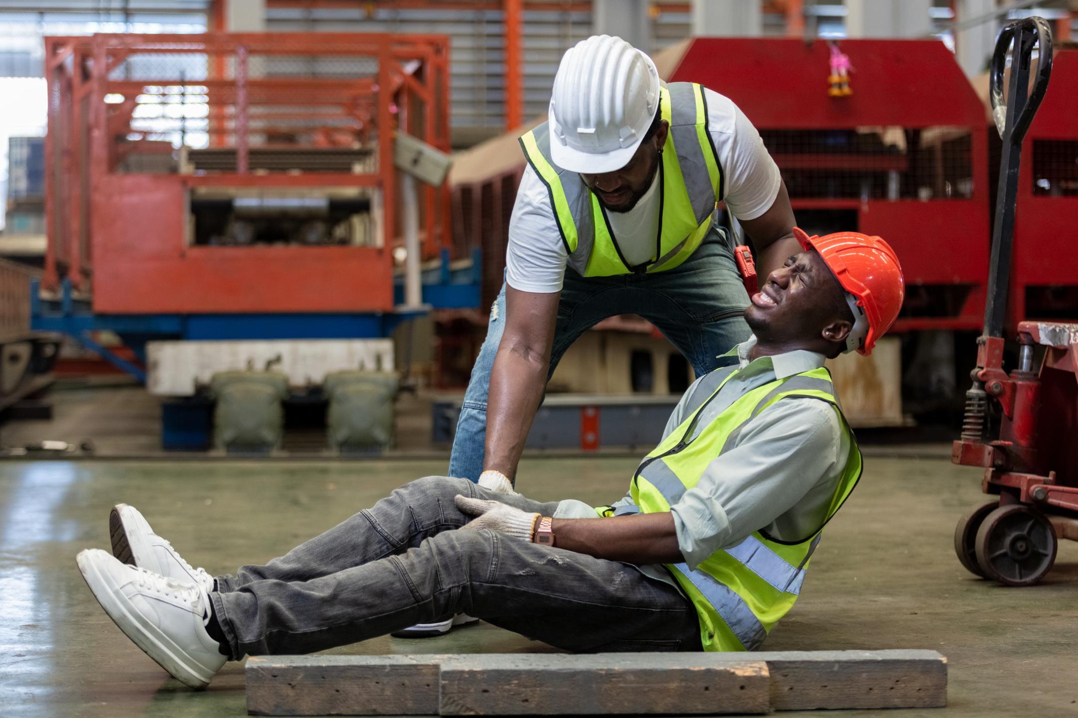 A worker in a safety vest and hard hat sits on the floor in pain while another worker assists him in an industrial setting.