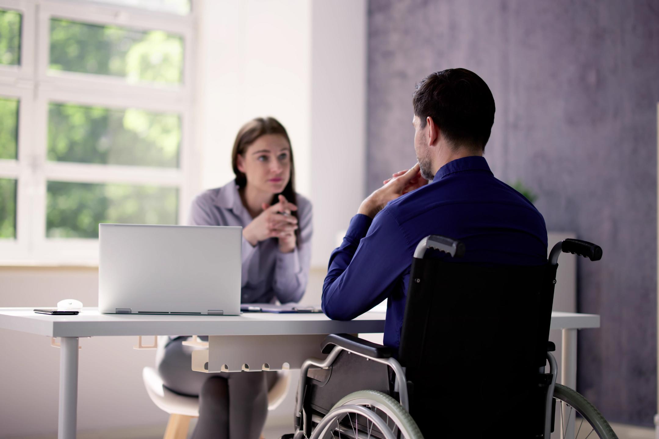 A man in a wheelchair discusses his case with a workers compensation lawyer in a professional office setting with a laptop and documents.