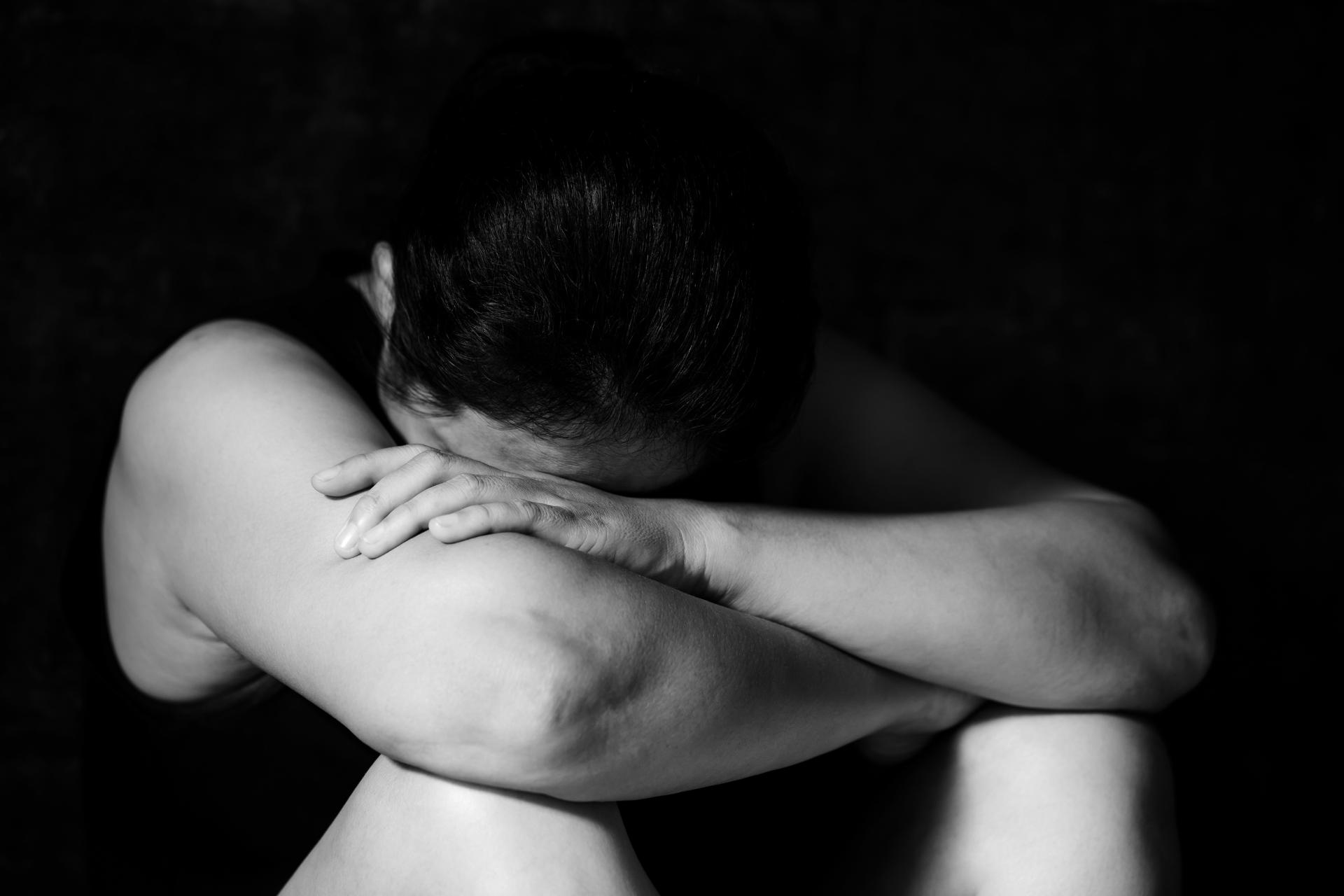 Person sitting with head down on crossed arms, knees drawn up, in a dark setting. The posture suggests sadness, distress, or contemplation—emotions often faced after trauma. An Assault Injury Lawyer can help support those through difficult times.
