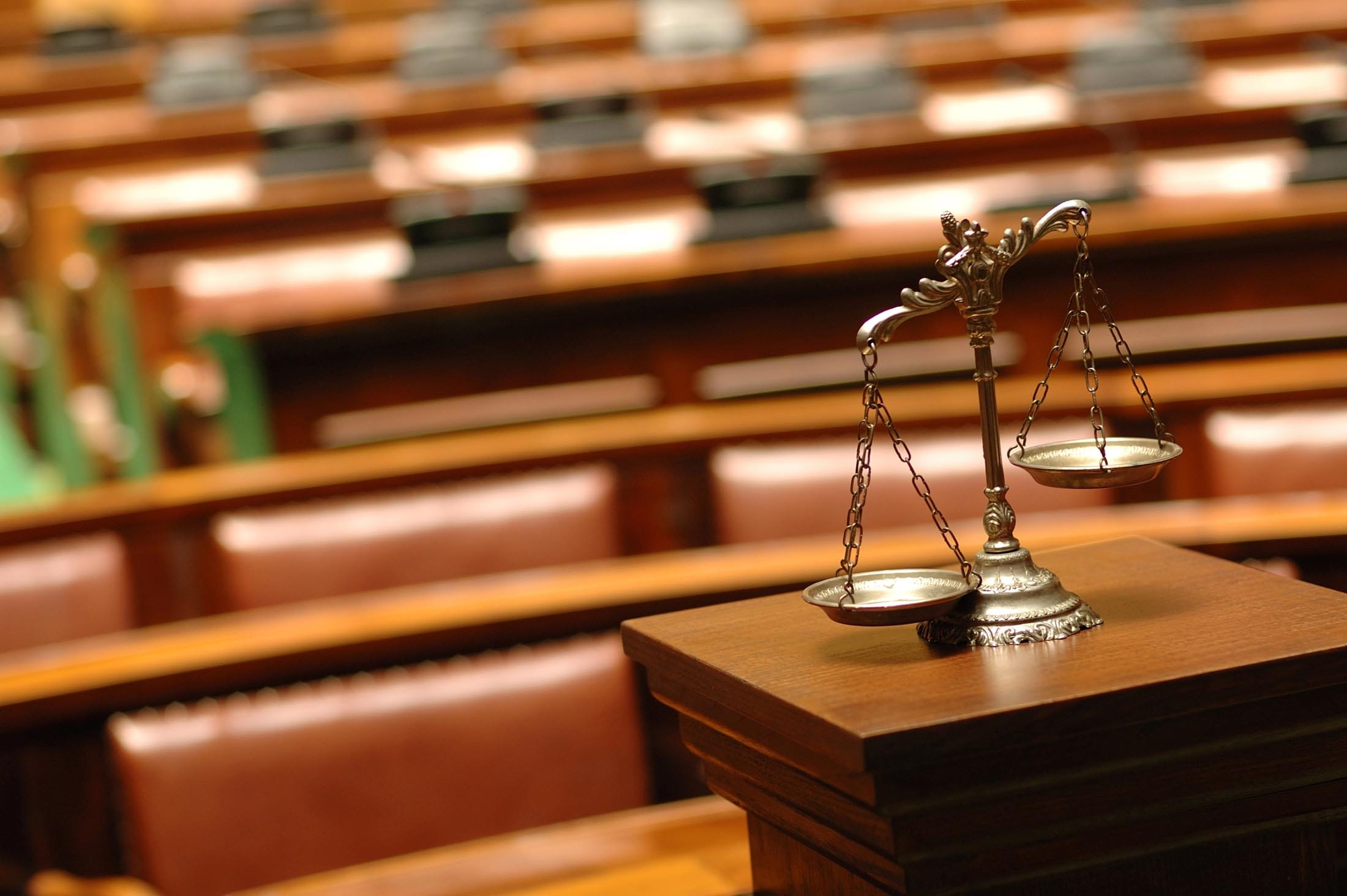 A brass balance scale sits on a wooden podium in an empty courtroom, with rows of brown leather seats in the background&mdash;a solemn scene reminiscent of justice and tradition, much like a classic Hummel Footer sculpture.