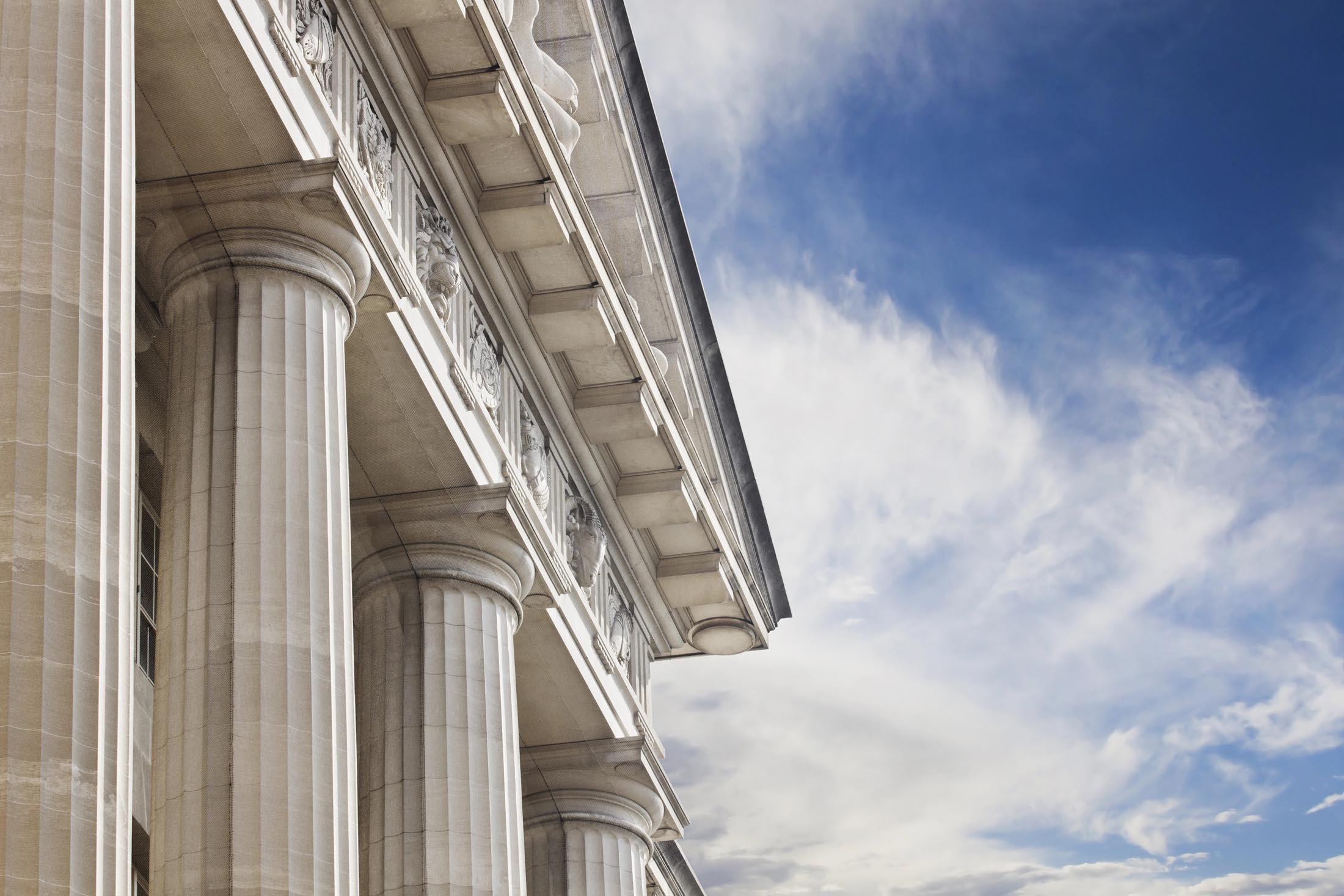 Close-up view of tall stone columns on a neoclassical building against a blue sky with scattered clouds, featuring intricate Hummel-inspired details at the column footer.
