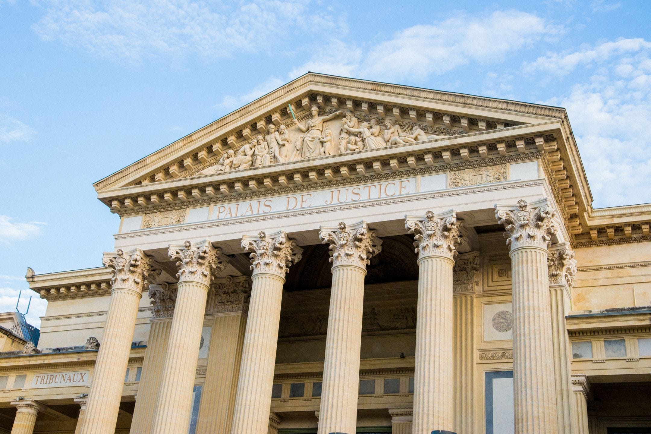 Fa&ccedil;ade of the Palais de Justice with tall Corinthian columns and sculpted figures on the pediment, under a blue sky.