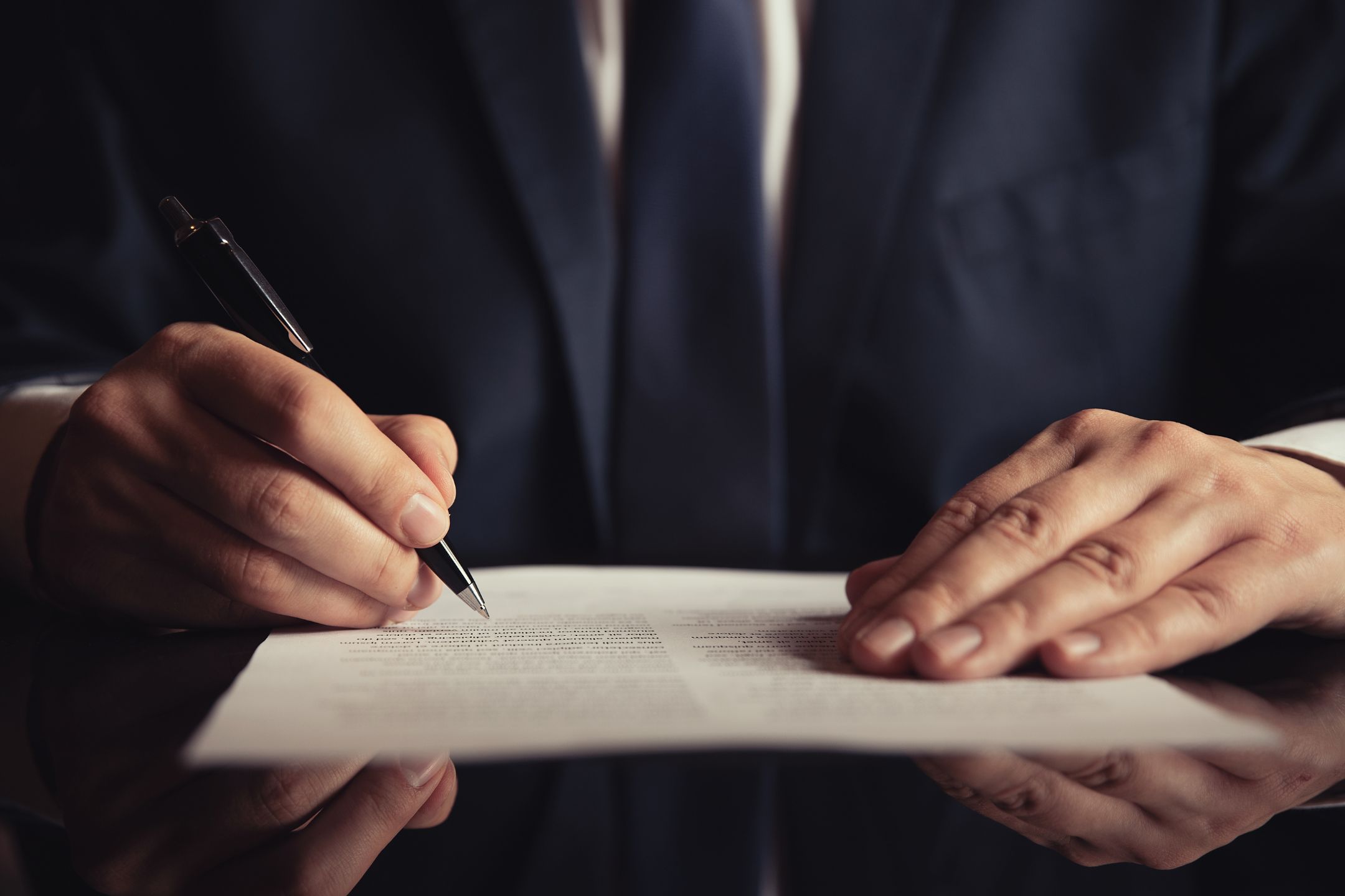 A person in a dark suit signs a document with a pen at a desk, with only their hands and torso visible.