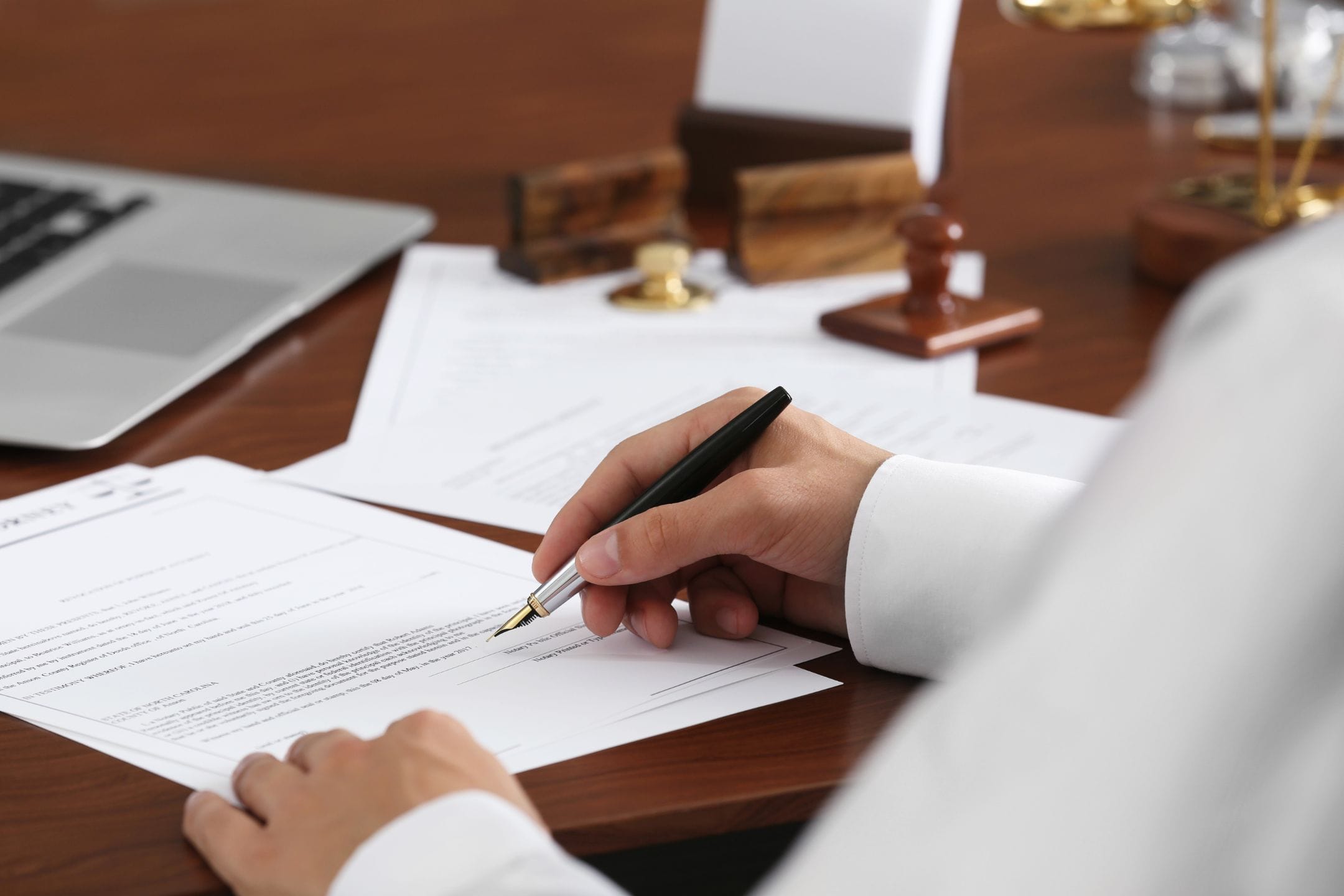 A person in formal attire signs a document at a desk with legal items, papers, a laptop, and a scale of justice visible.