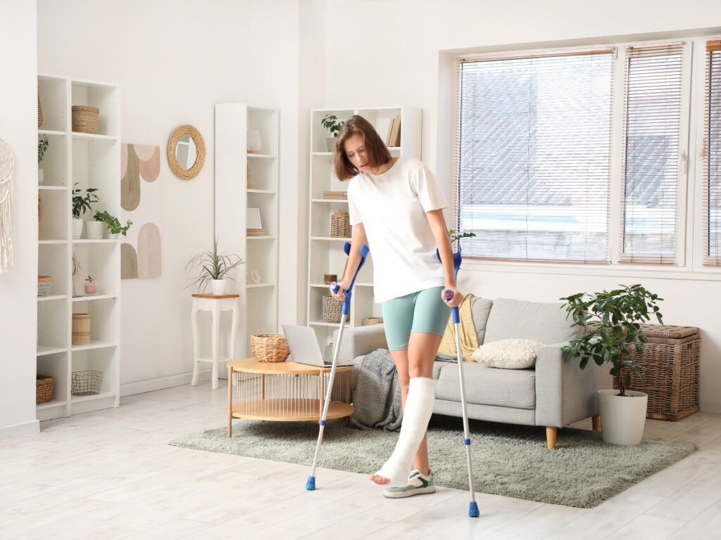 A woman with a cast on her right leg, recovering from one of the common slip and fall injuries, uses crutches to walk in a bright, modern living room with plants and neutral decor.