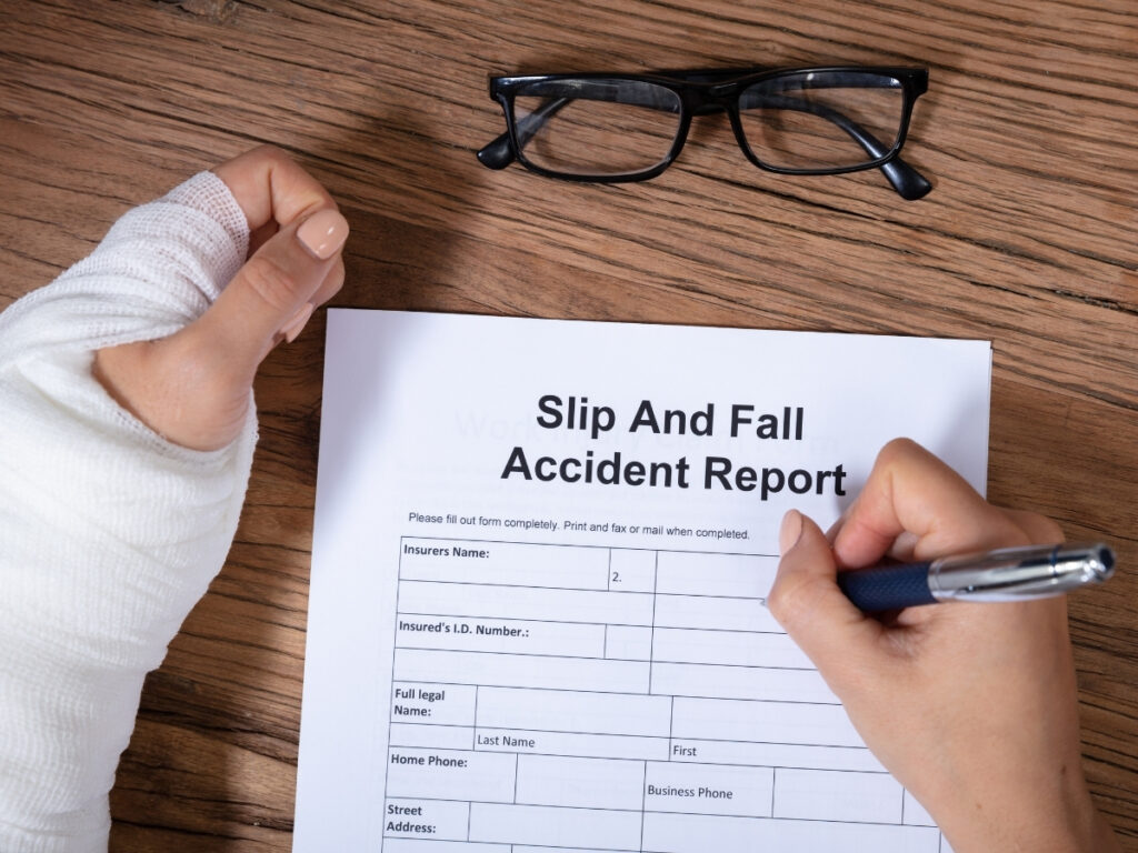 A person with a bandaged arm, showing signs of common slip and fall injuries, fills out an accident report form on a wooden table, with a pair of glasses nearby.