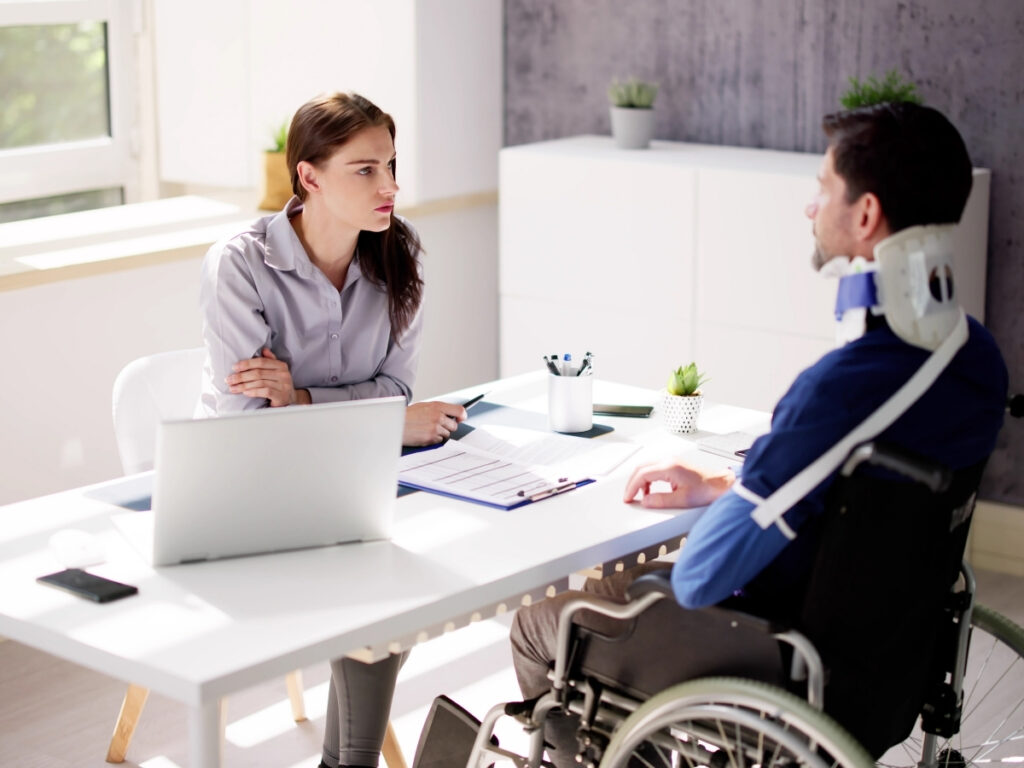 A woman sits at a desk with a laptop and paperwork, discussing how insurance companies evaluate personal injury claims with a man in a wheelchair who is wearing a neck brace.
