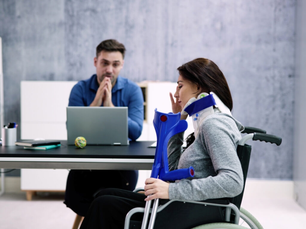 A woman in a wheelchair with a neck brace and crutches sits across a desk from a man using a laptop, discussing how insurance companies evaluate personal injury claims in an office setting.