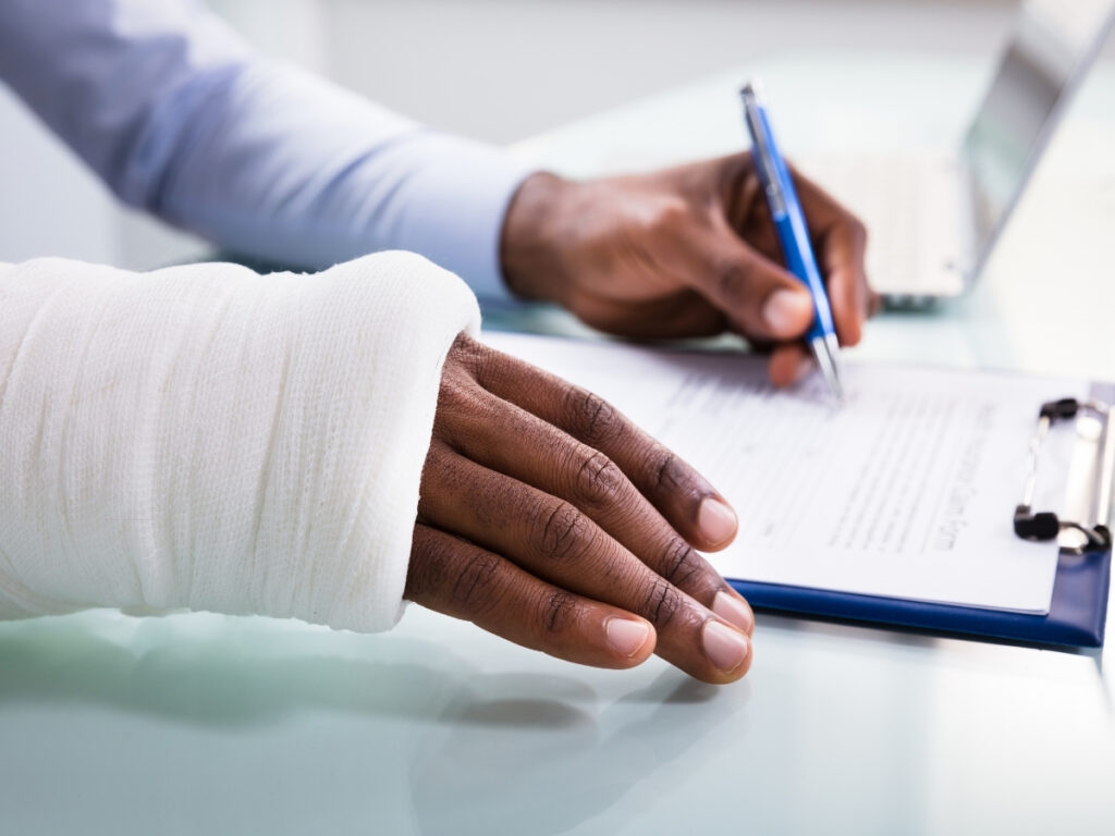 A person with a bandaged arm is filling out a form on a clipboard, possibly documenting details relevant to how insurance companies evaluate personal injury claims, seated at a desk with a laptop nearby.
