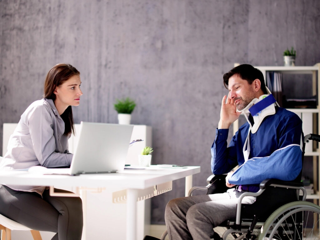 A woman sits at a desk with a laptop, listening to a man in a wheelchair wearing a neck brace and arm sling discuss how insurance companies evaluate personal injury claims in an office setting.