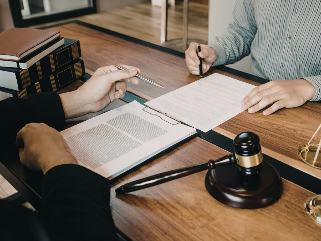 Two people sit at a desk with legal documents, a gavel, books, and a scale; one person is signing a paper while the other explains how insurance companies evaluate personal injury claims.