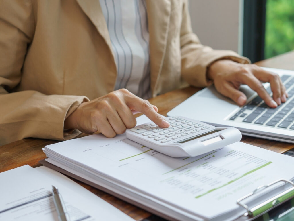 Person in a beige blazer using a calculator and laptop, working with financial documents on a wooden desk—researching how insurance companies evaluate personal injury claims.