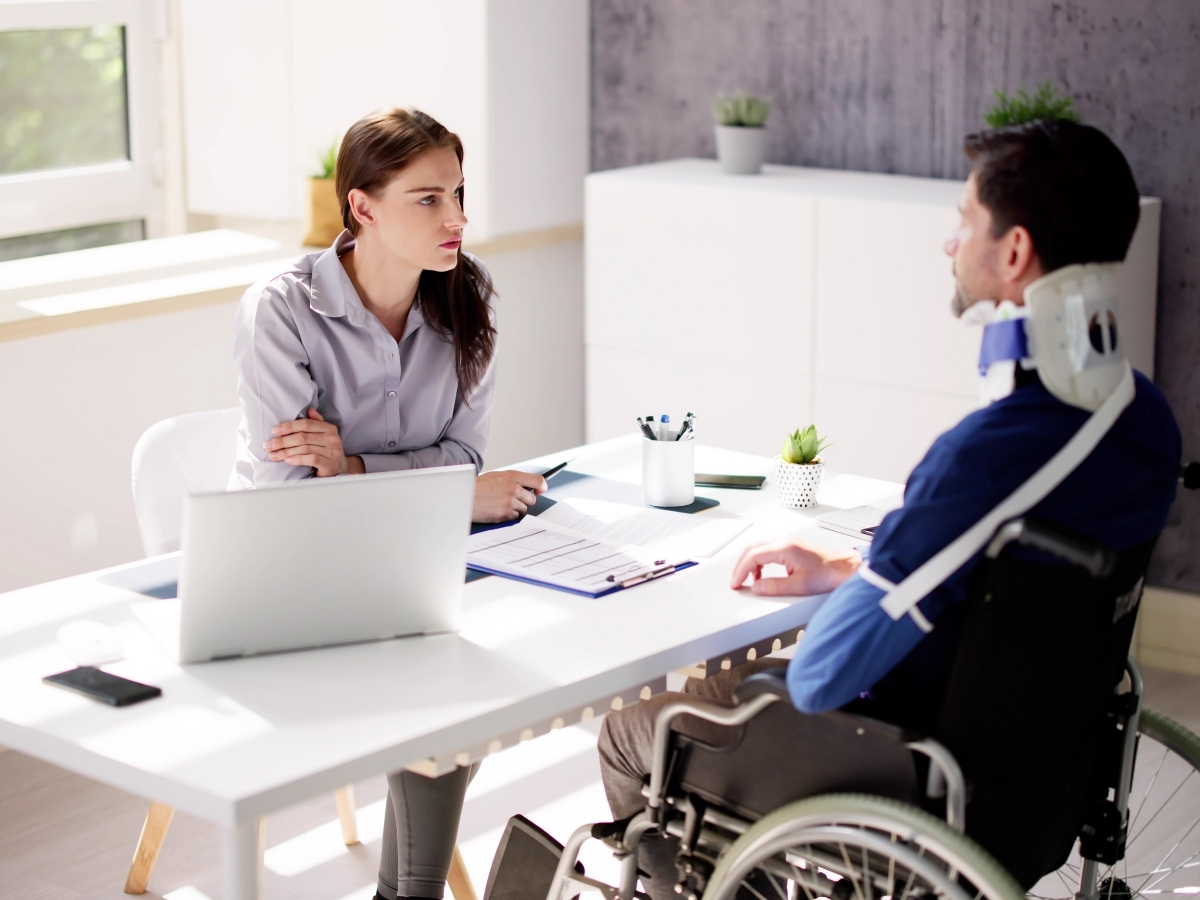 A woman sits at a desk with a laptop and paperwork, discussing how insurance companies evaluate personal injury claims with a man in a wheelchair who is wearing a neck brace.