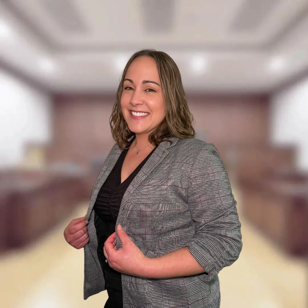 A woman in a plaid blazer stands smiling in a blurred indoor setting resembling a courtroom or office.