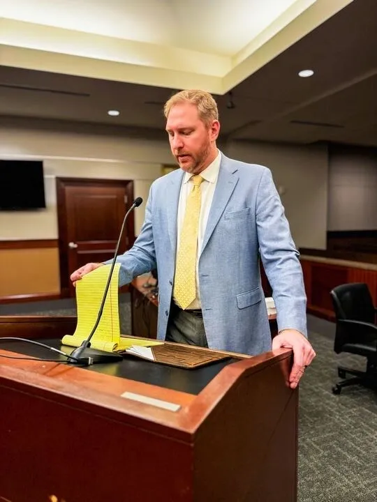 A man in a light blue suit stands at a podium in a courtroom, holding a yellow notepad and speaking into a microphone.