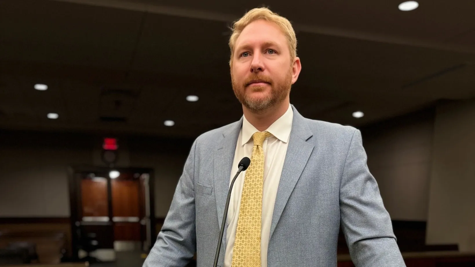 A man in a light gray suit and yellow tie speaks at a podium, an image featured in our attorney bio to highlight his history of courtroom advocacy.