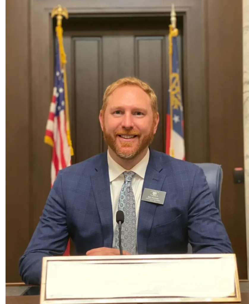 A man in a blue suit sits at a desk with a nameplate, smiling, with two flags and a wooden door in the background.
