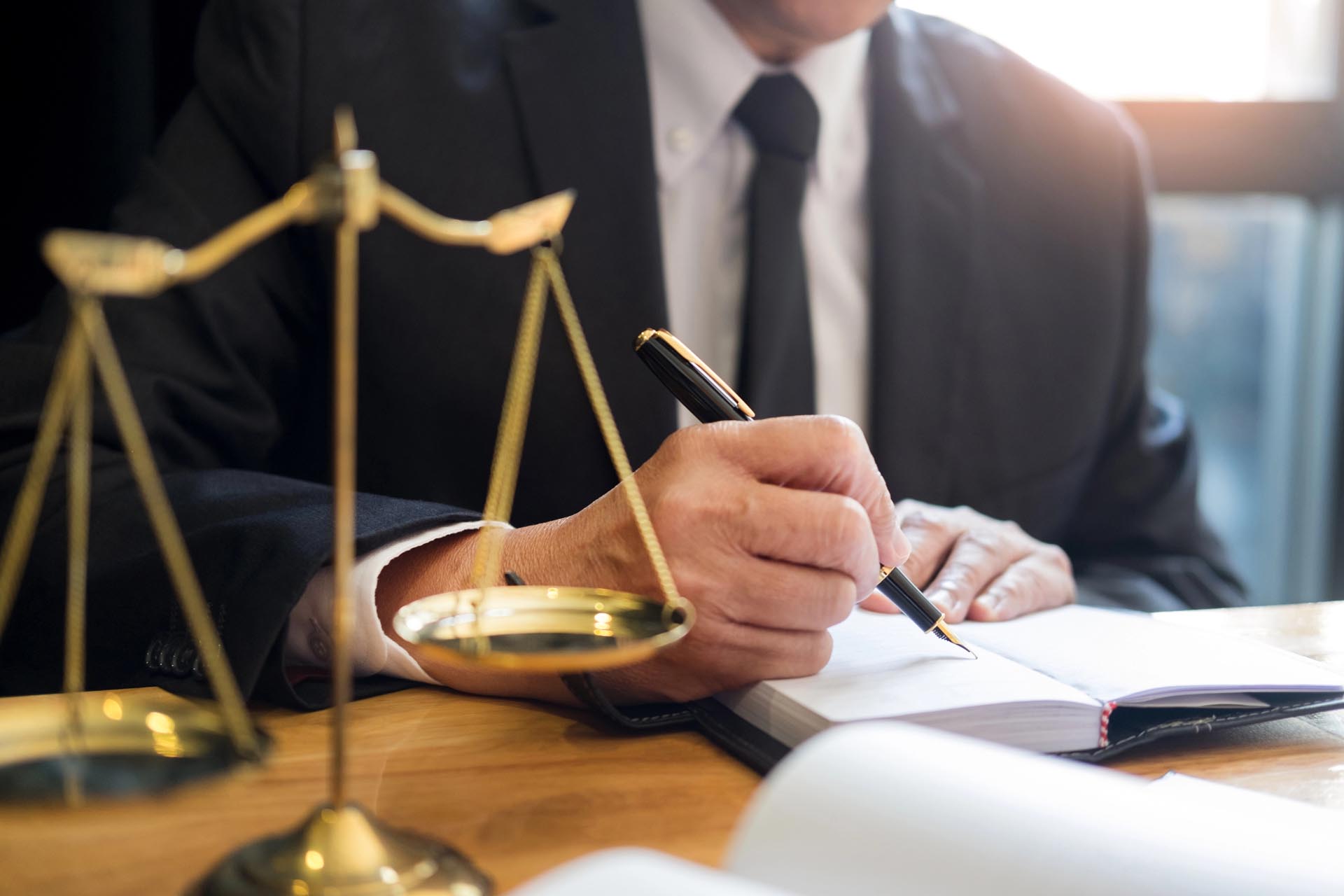 A person in a business suit writes in a notebook at a desk, with a balance scale symbolizing law or justice in the foreground, highlighting various legal practice areas.