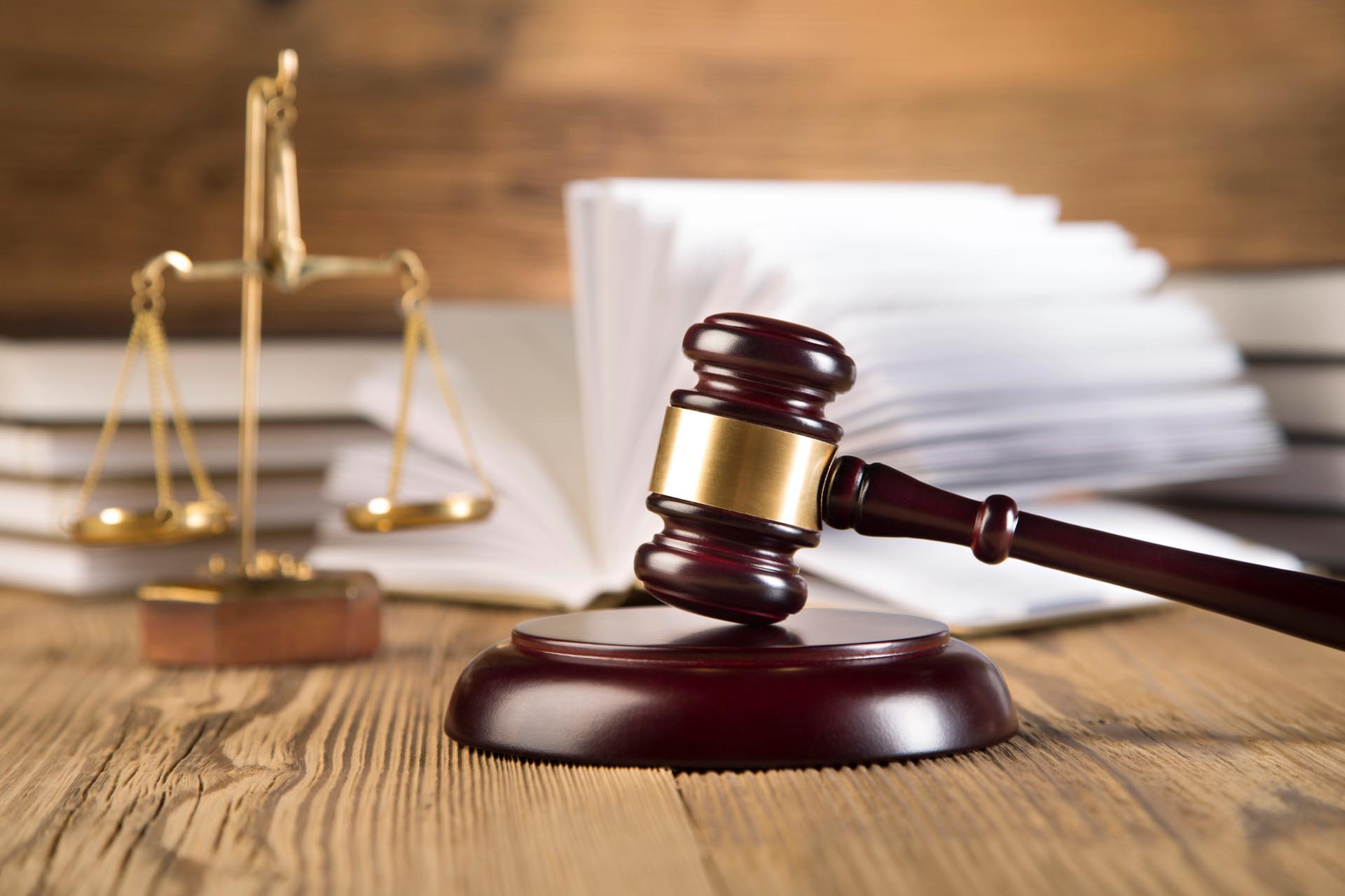A wooden gavel rests on a sound block beside brass scales of justice and an open book, symbolizing diverse practice areas, atop a wooden table.