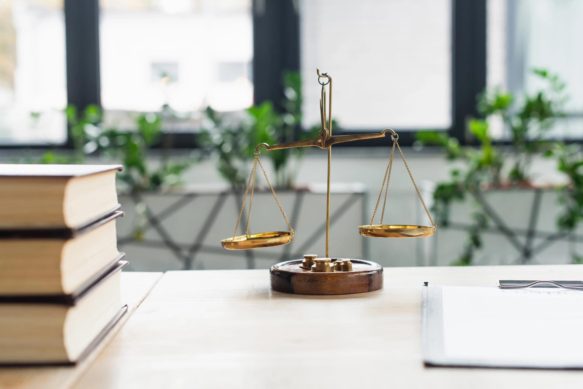 A brass balance scale sits on a desk next to a stack of books, symbolizing diverse practice areas, with greenery visible outside large windows in the background.
