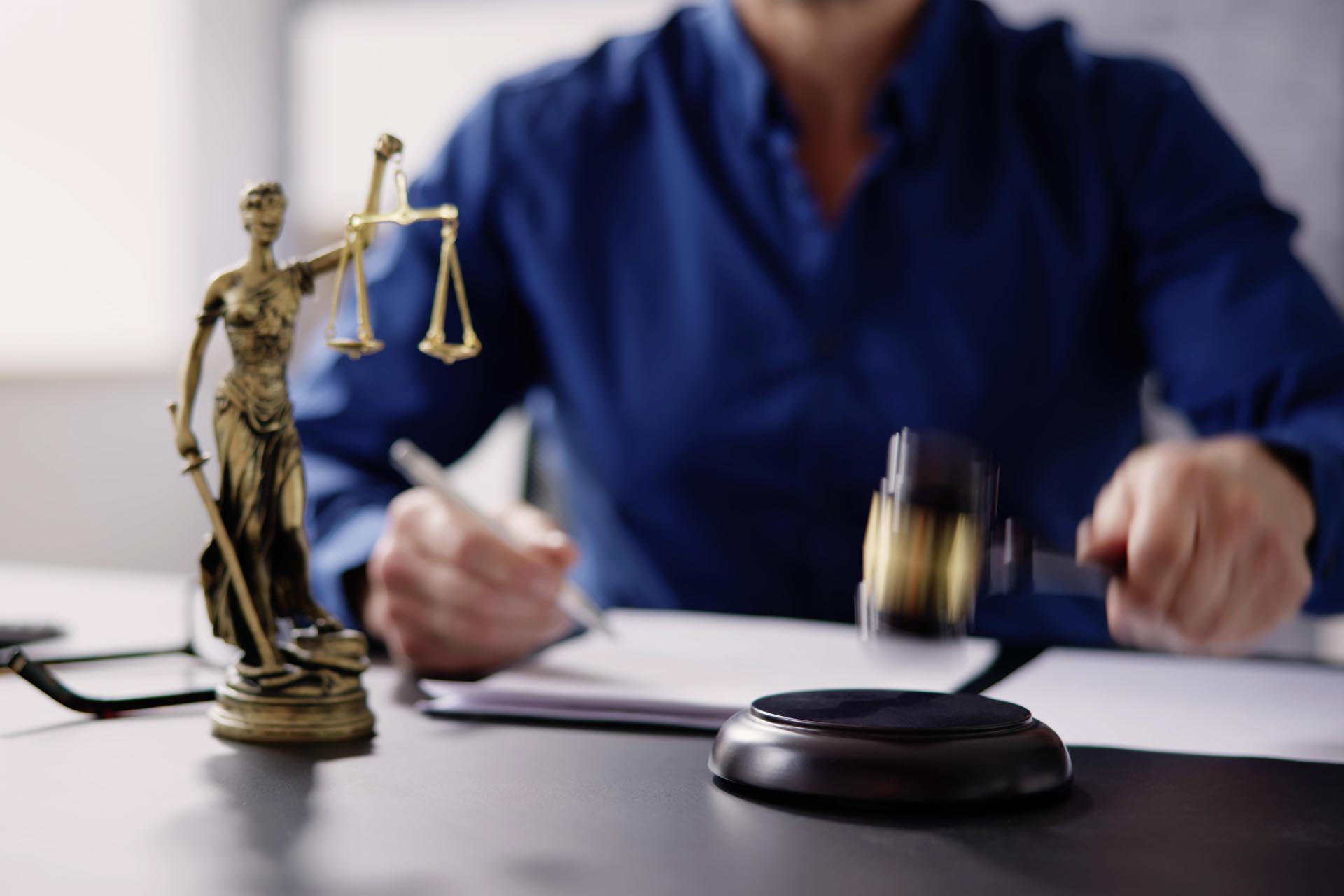 A person in a blue shirt uses a gavel at a desk with documents, a small statue of Lady Justice, and information about practice areas.