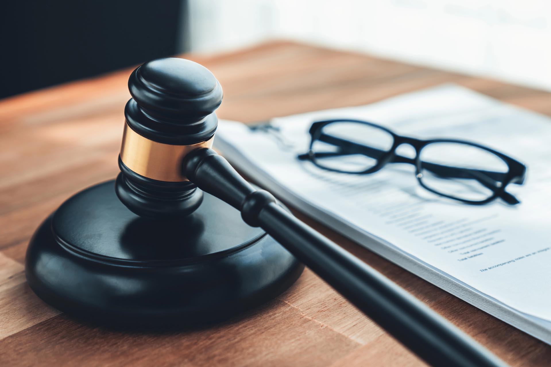 A judge’s gavel rests on a wooden table next to a stack of papers detailing various practice areas and a pair of black eyeglasses.