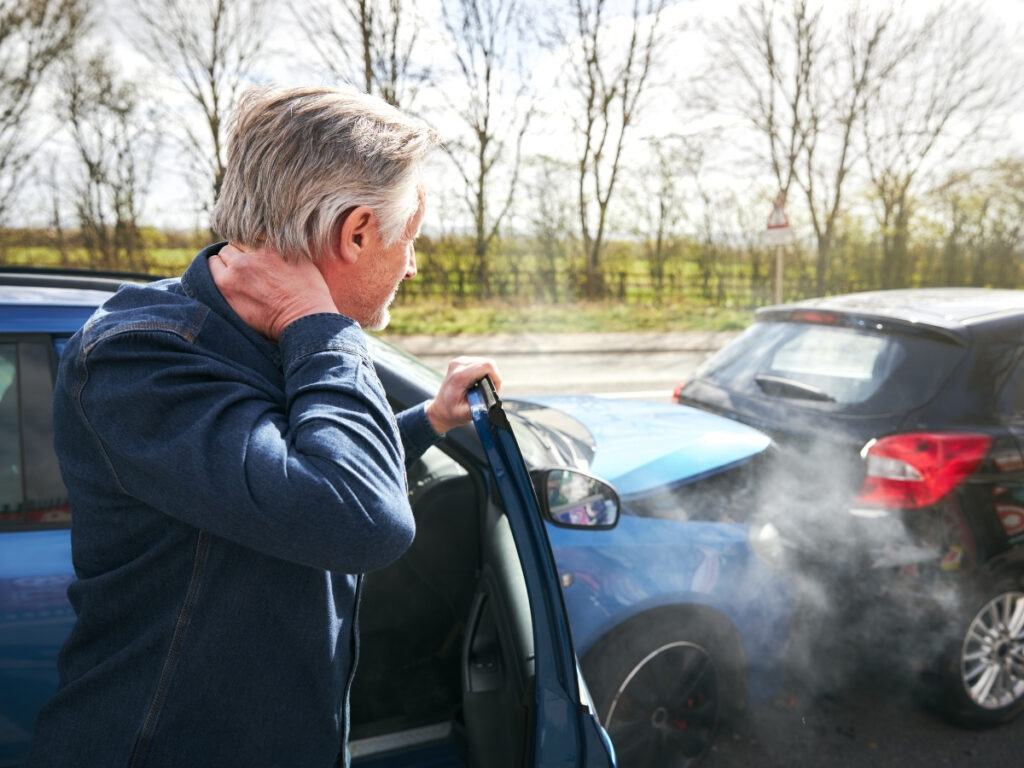 A man holding his neck stands beside his car after a rear-end collision with another vehicle; smoke is visible from the front of his car, illustrating what to do after a car accident in Georgia.