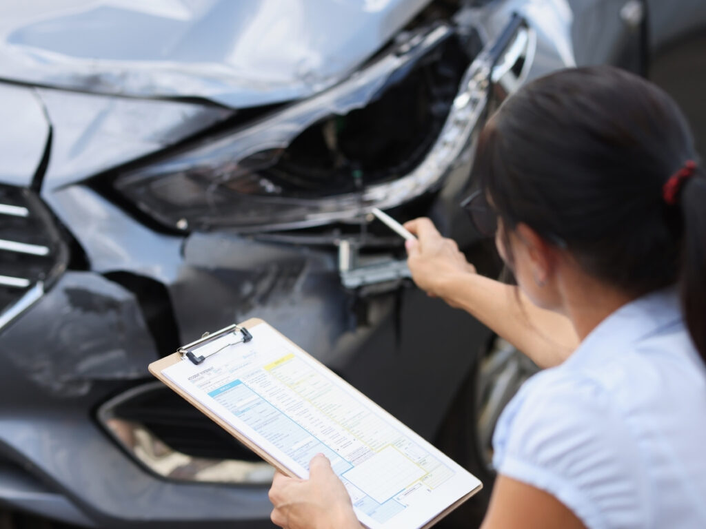 A person holding a clipboard inspects and points at damage on the front of a gray car, demonstrating what to do after a car accident in Georgia.