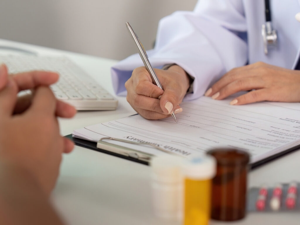 A doctor fills out a medical form while a patient sits across the desk, possibly discussing what to do after a car accident in Georgia; medication bottles and a keyboard are visible on the table.