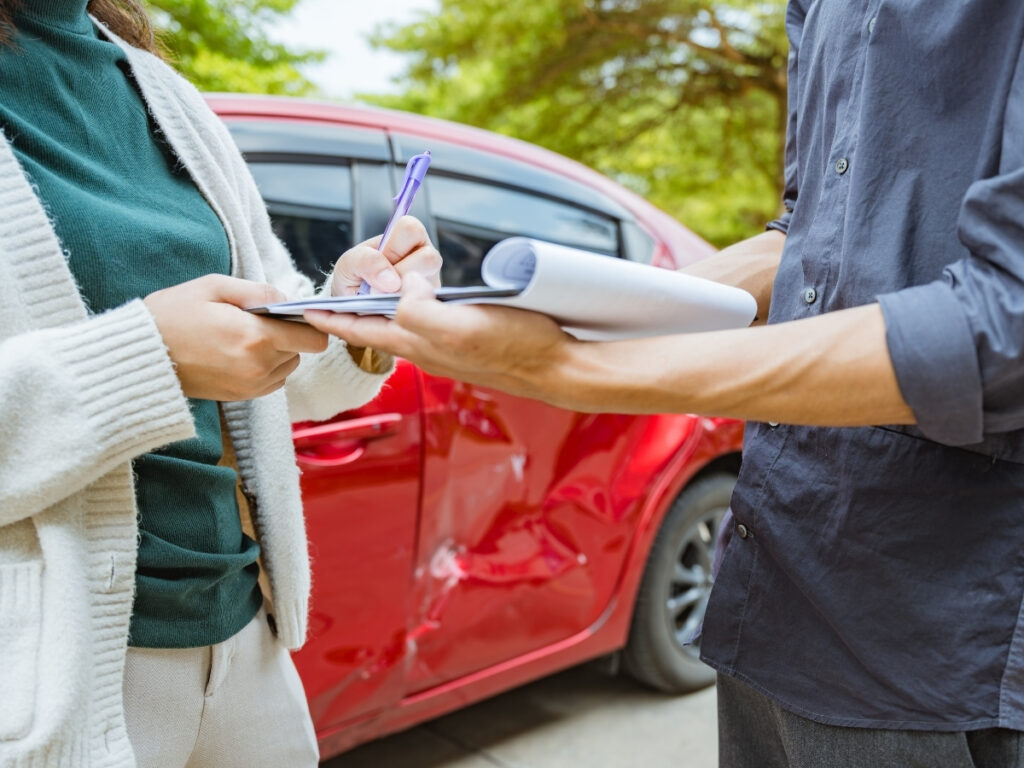 Two people stand beside a red car with a damaged door, exchanging information and writing on a clipboard and notepad—an important step in what to do after a car accident in Georgia.