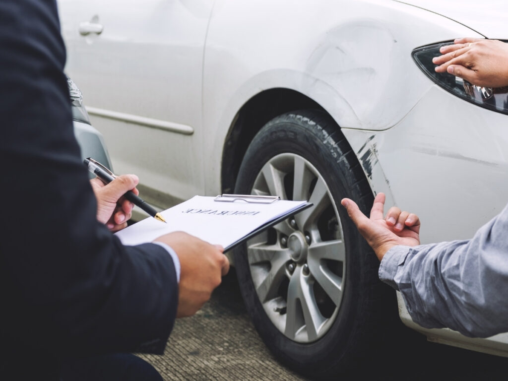 Two people inspecting scratches on a white car’s front fender; one holds an insurance claims clipboard and pen, discussing what to do after a car accident in Georgia.