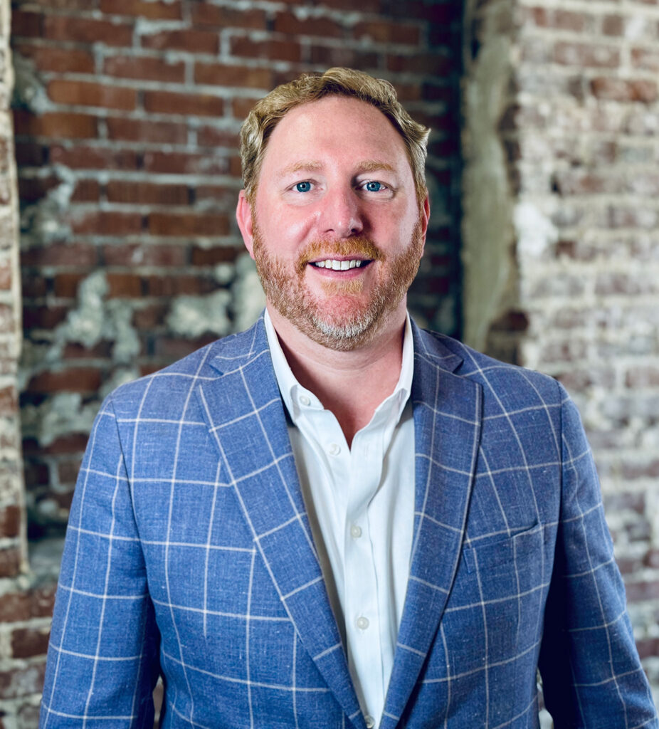 A man with light brown hair and a beard, wearing a blue plaid blazer and white shirt, stands in front of a weathered brick wall, smiling warmly at the camera as if about to share his story.