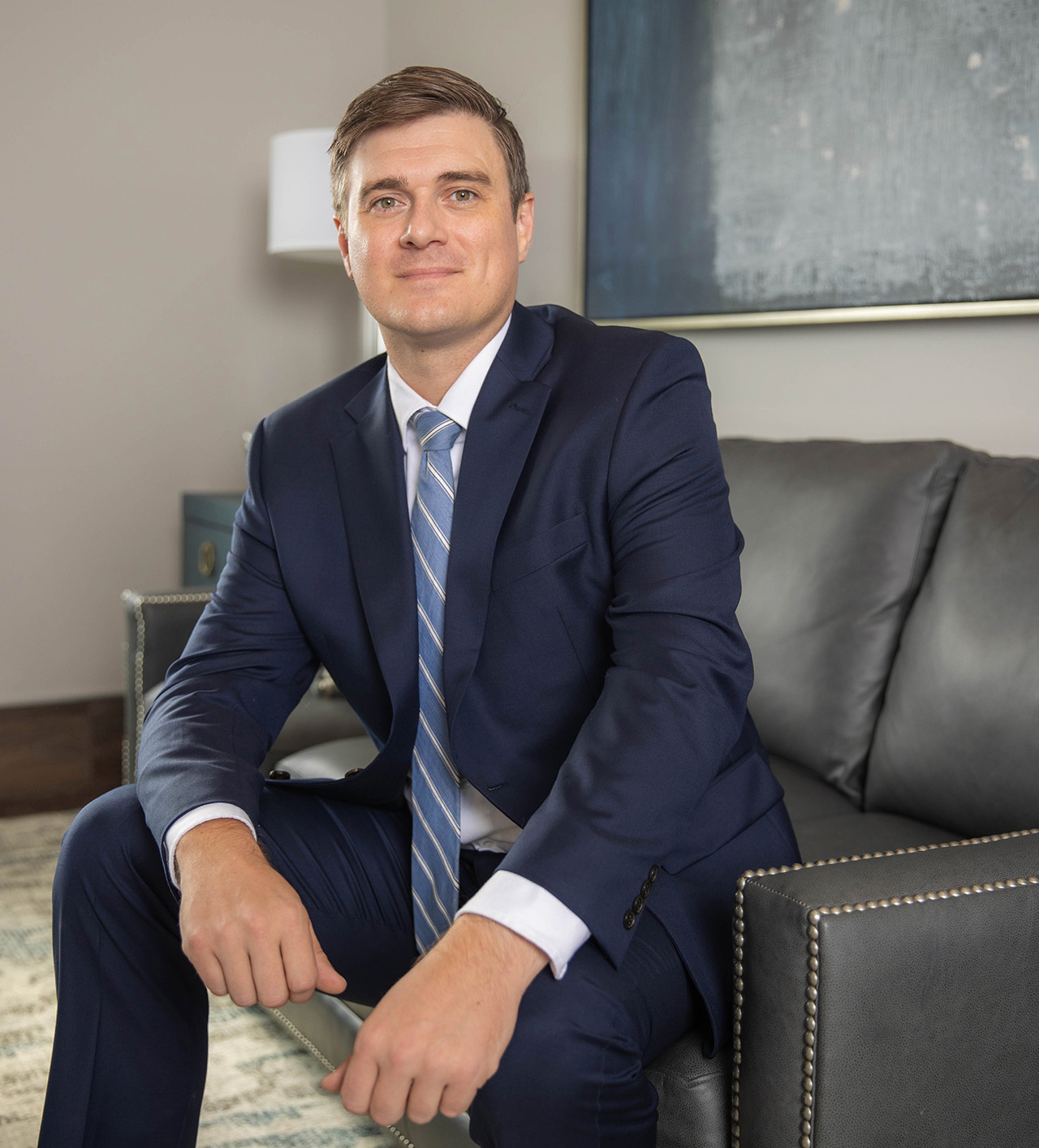 A man in a blue suit and striped tie sits on a gray couch, looking about the room with a neutral expression. A painting and side table are visible in the background.