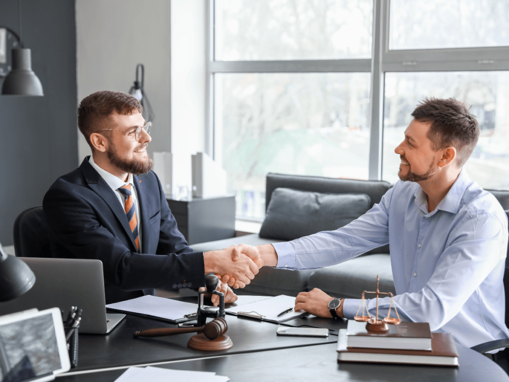 Two men, one in a suit and one in a dress shirt, shake hands across a desk in an office with legal symbols and documents visible, representing the trusted relationship between client and Georgia personal injury lawyer.