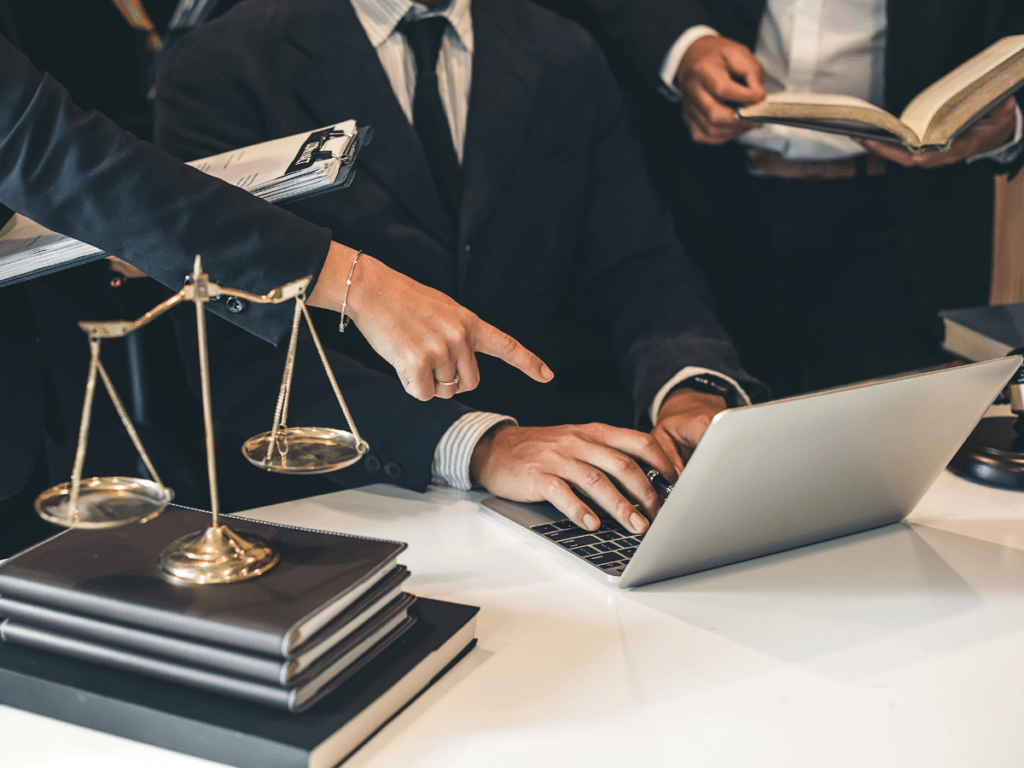 Three people in business attire work at a desk with a laptop, law books, and scales of justice; one points at the screen while another holds an open book, demonstrating the collaborative approach of a Georgia Personal Injury Lawyer providing skilled representation to their clients.