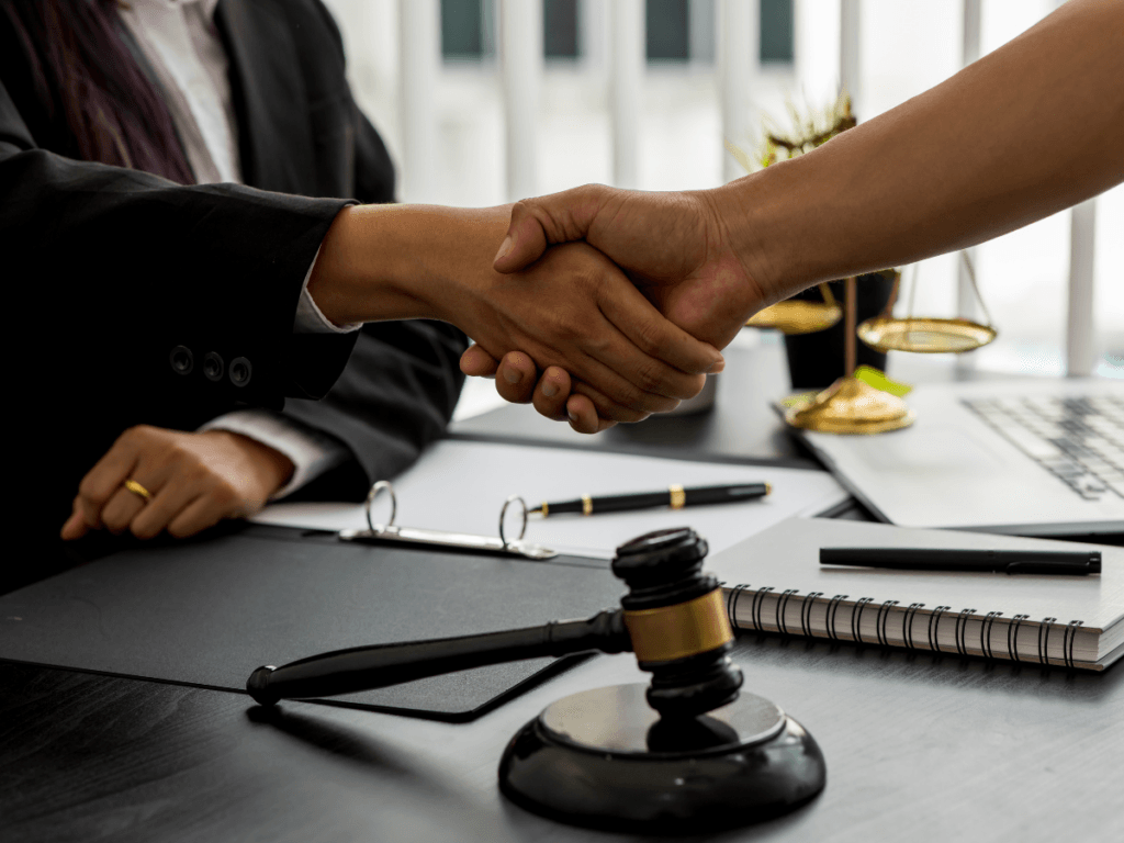 Two people shake hands across a desk with a gavel, a legal document, and scales of justice, suggesting a legal agreement or consultation with a Personal Injury Lawyer Georgia.