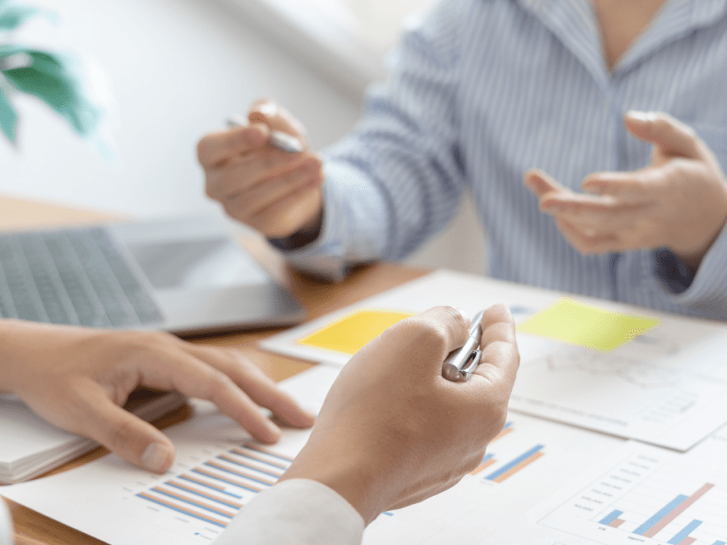 Two people in business attire discuss graphs and charts at a desk, holding pens, as they evaluate settlement options. A laptop and documents are visible, emphasizing collaborative decision making.