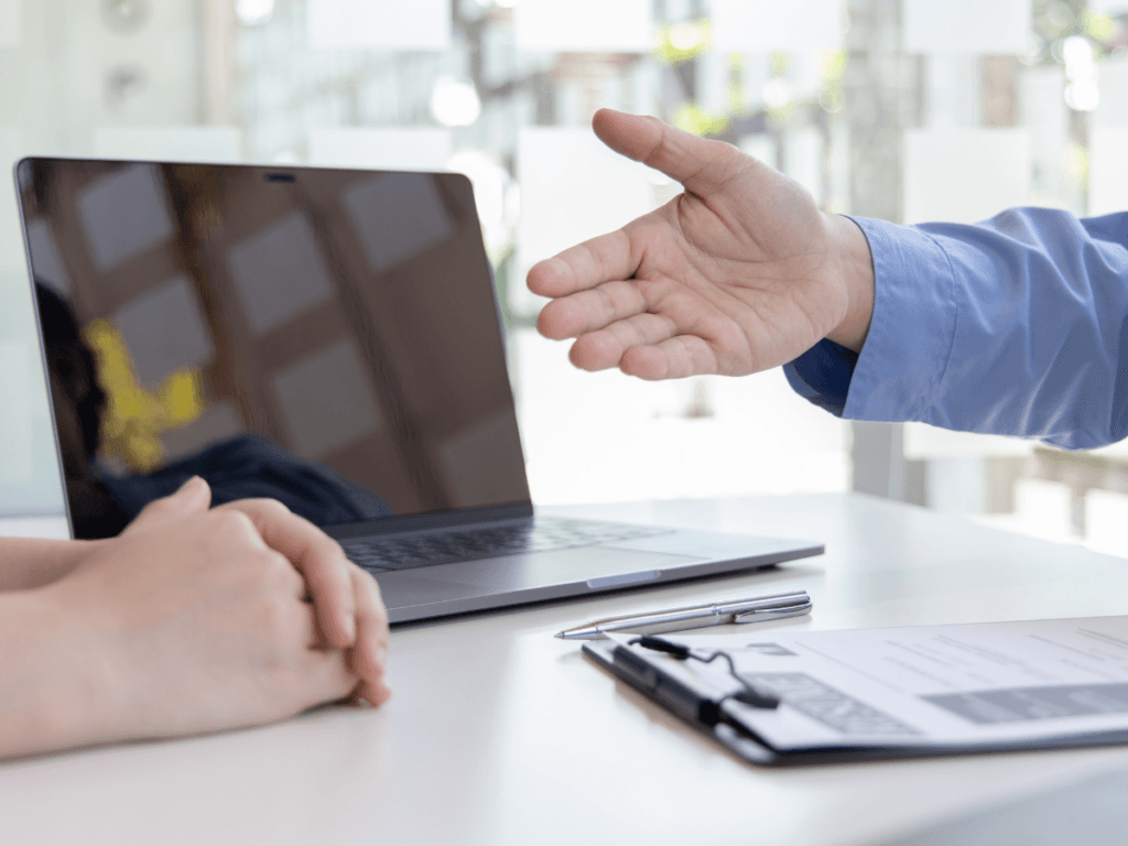 Two people sit at a desk with a laptop, pen, and clipboard; one person gestures with their hand while the other listens closely, as they evaluate settlement options during the decision-making process.