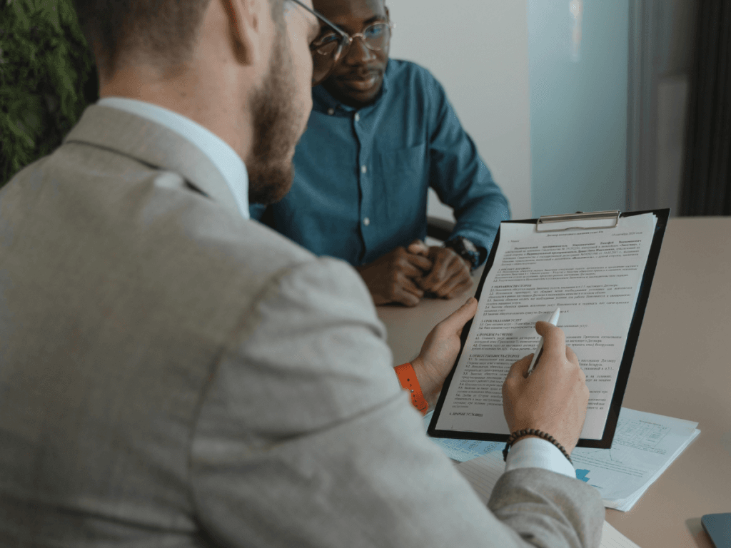 Two people sit at a table; one holds a clipboard with paperwork and points at a section with a pen while the other person listens attentively, carefully considering how to evaluate settlement options before making a final decision.