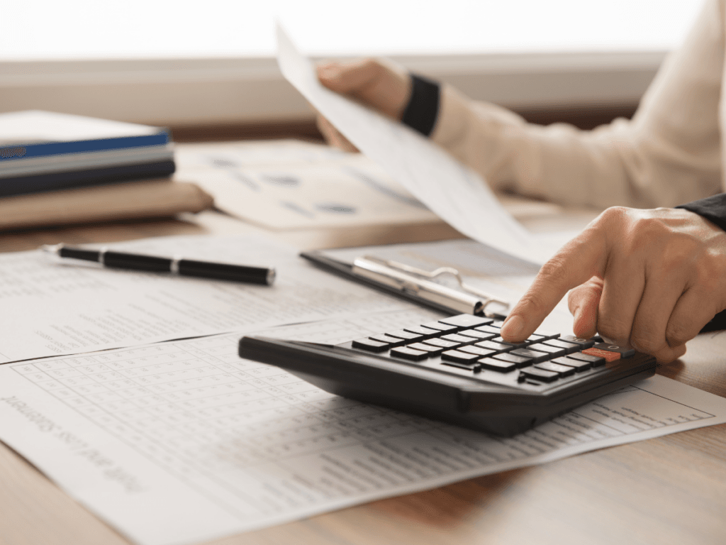 Person engaged in financial planning, using a calculator and holding a paper at a desk with financial documents, a pen, and a clipboard.