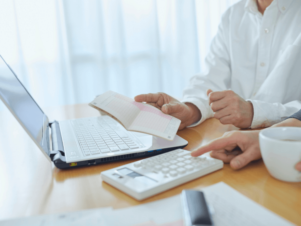 Two people review a bankbook and use a calculator in front of a laptop on a wooden table, with documents and a cup nearby, as part of their careful financial planning.