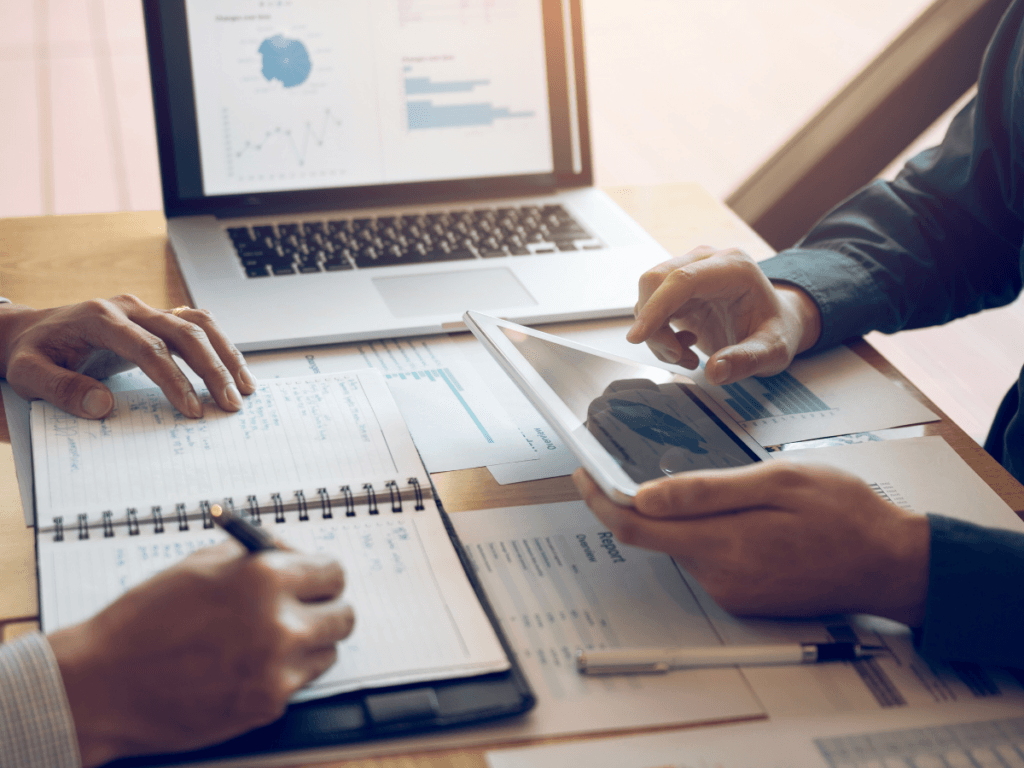 Two people review documents and notes at a desk with financial charts on a laptop and tablet, discussing data, financial planning, and writing in a notebook.