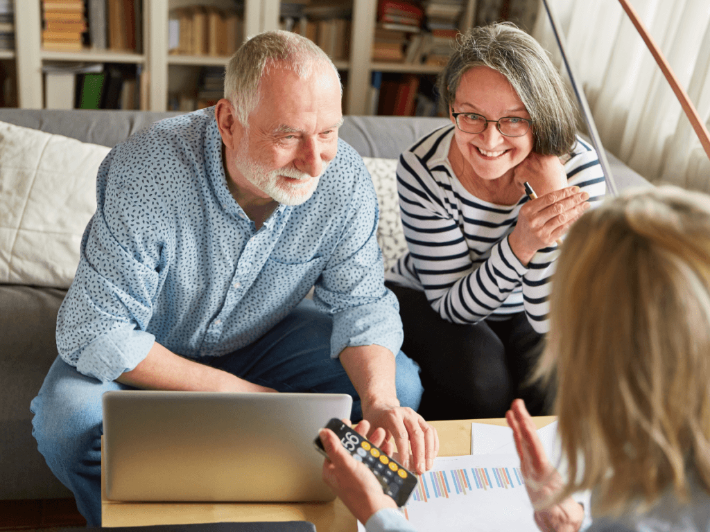 Two older adults sit on a couch, smiling and looking at a laptop, engaged in financial planning. Another person holds a calculator and gestures toward papers with charts on a table.