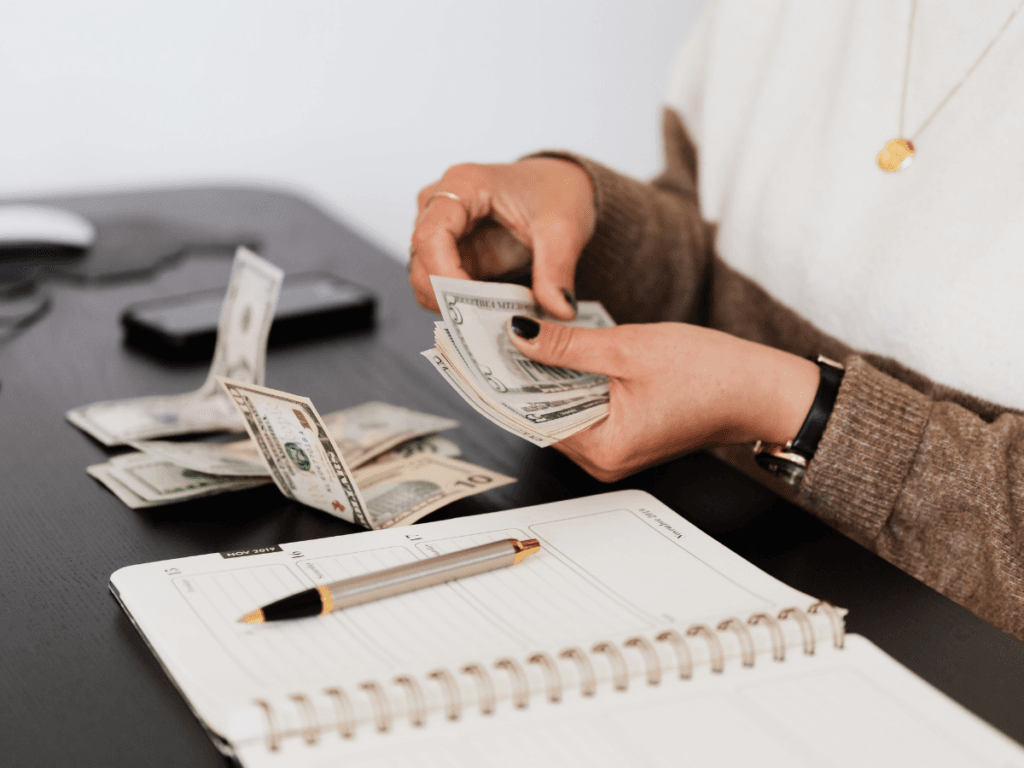 A person counts US dollar bills at a desk with a pen and open notebook in front of them, carefully engaged in financial planning.