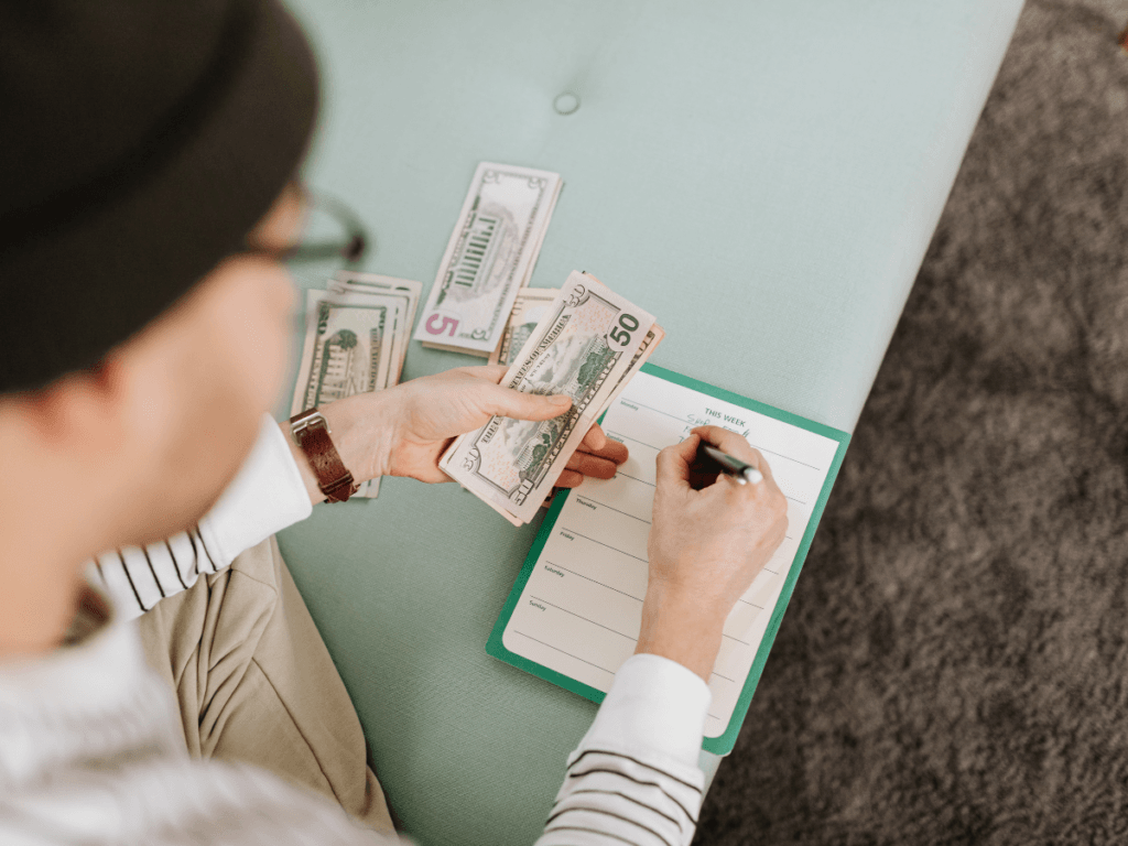 A person holding US dollar bills writes on a notepad while sitting on a light green sofa, engaged in financial planning.
