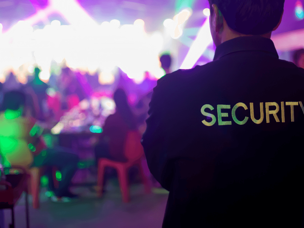 A security guard stands in the foreground at a crowded nightclub event in Georgia, with colorful lights and people sitting at tables in the background—a reminder of the importance of preventing negligent security incidents.