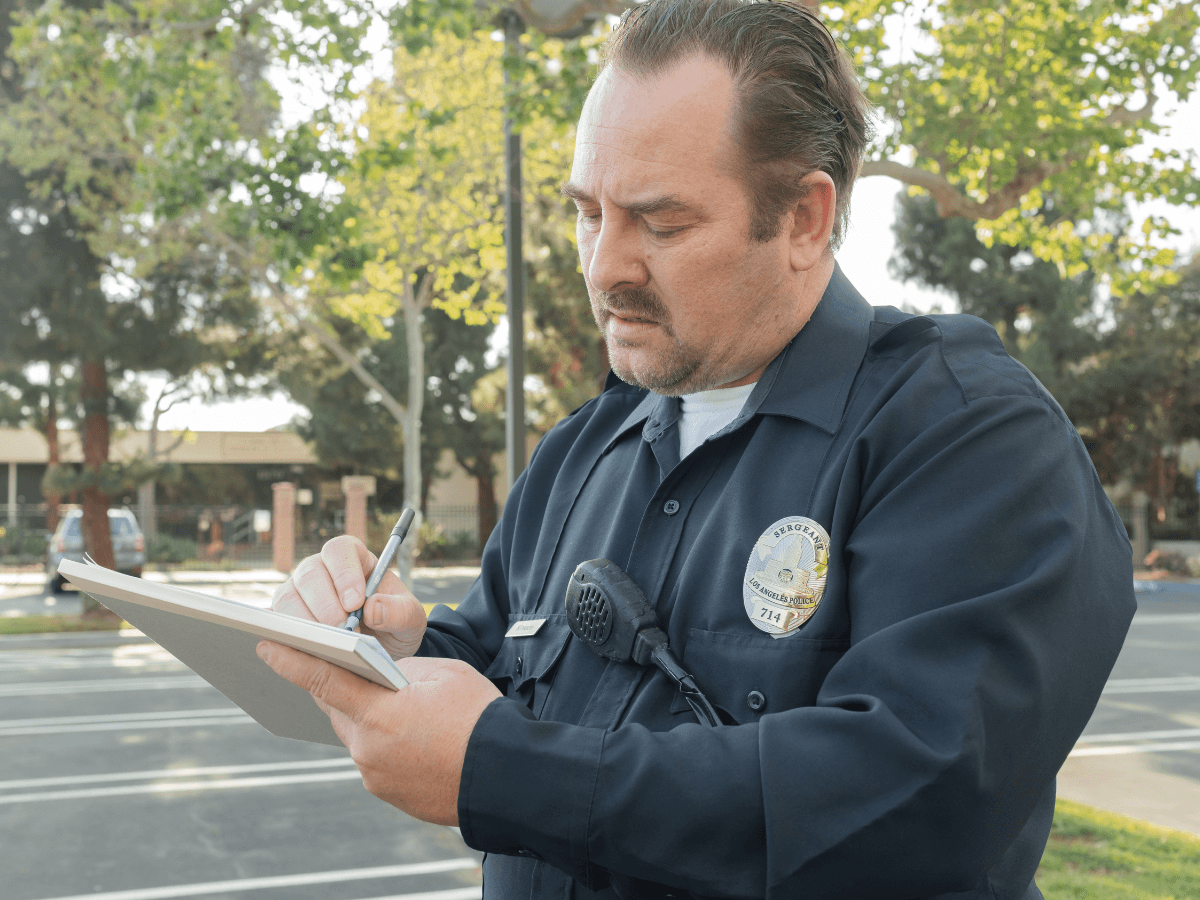 A police officer stands outdoors, looking down while writing on a notepad, carefully documenting details to avoid police report errors. Trees and a building are visible in the background.