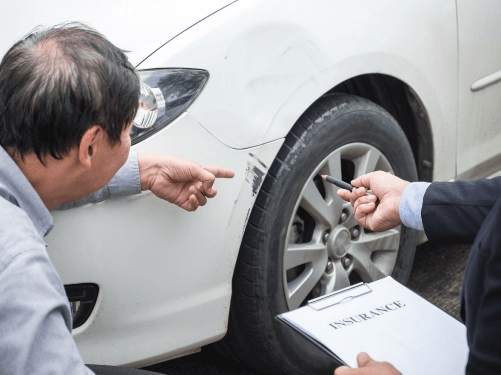 Two people inspect scratches on a white car’s fender. One person points at the damage while the other holds a clipboard labeled “insurance” and a pen, carefully following their car crash checklist to ensure they complete all first steps after car crash.
