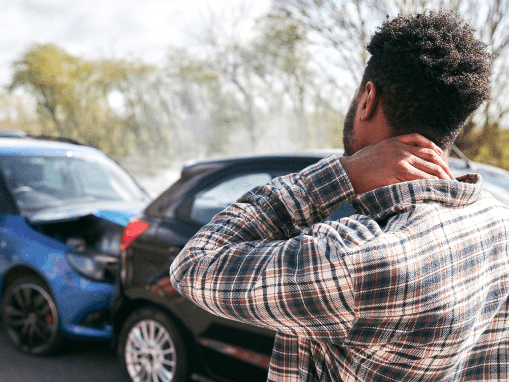 A man holds his neck while standing near two cars involved in a rear-end collision on a road during the day, wondering about the first steps after car accident and referring to his car crash checklist.