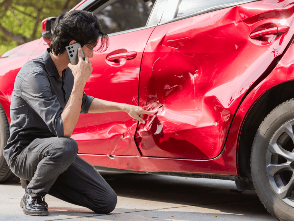 A person crouches next to a red car with a large dent on the door, speaking on a phone and inspecting the damage—clearly focused on what to do after a car crash and taking the essential first steps from their car crash checklist.